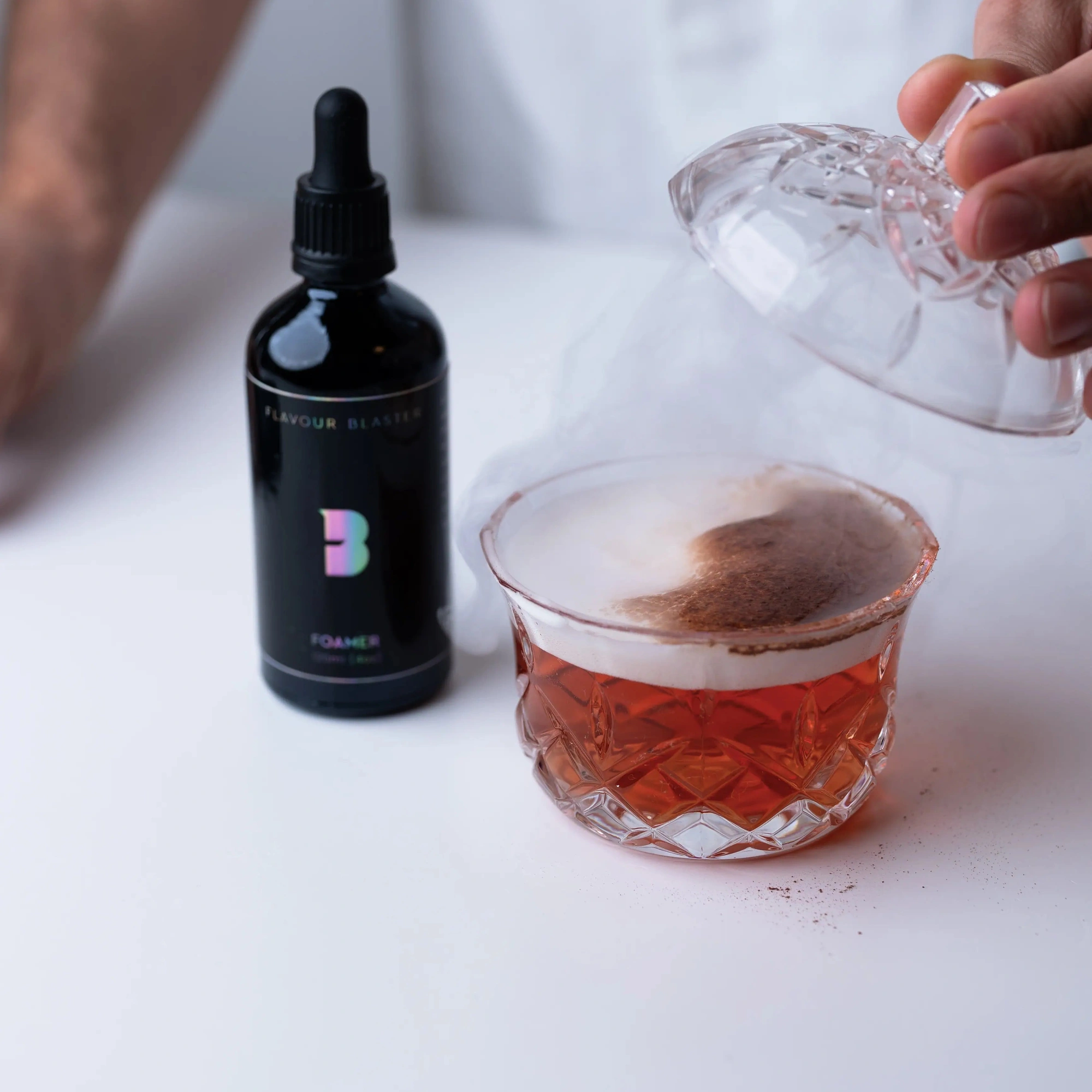 foamer cocktail Flavour Blaster: espuma sobre un cóctel rojo en un vaso de cristal tallado, con la campana de vidrio levantada y la botella al lado